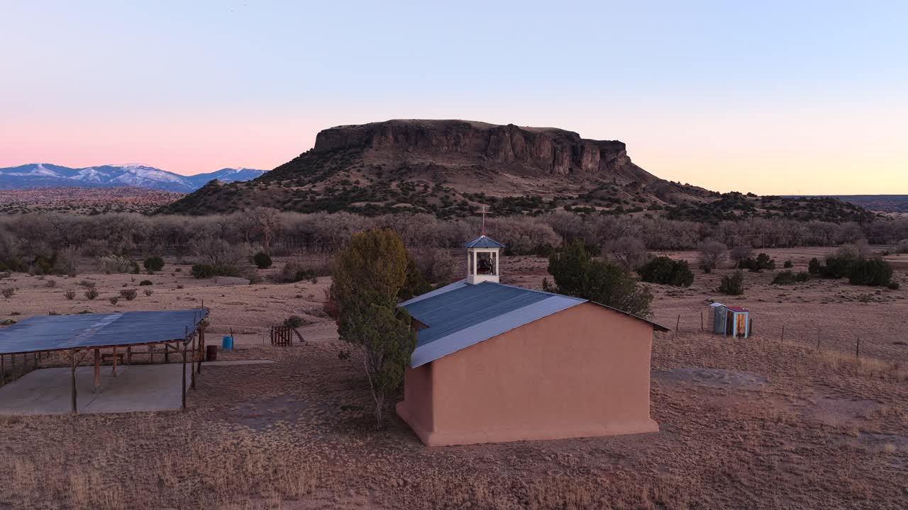 Plano geral de drone de alta qualidade se afasta e orbita a cruz de uma igreja rural ao entardecer. Revela uma vasta paisagem desértica com uma grande mesa e montanhas com picos nevados. Perfeito para aberturas de documentários