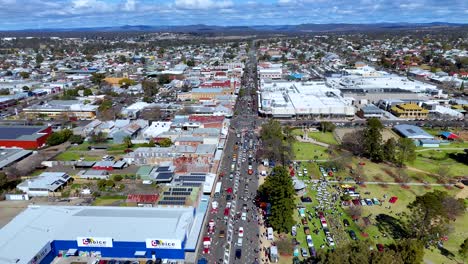 drone footage captures a vibrant street festival with crowds, classic cars, and market stalls along a main road in a small australian town under daylight