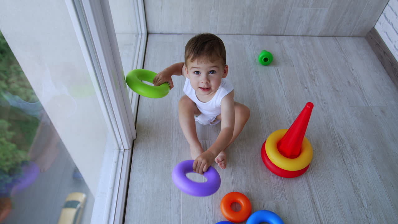 Cute kid in white bodysuit sits on the floor near panoramic window. Child plays with rings from toy pyramid banging them by the floor. Top view.