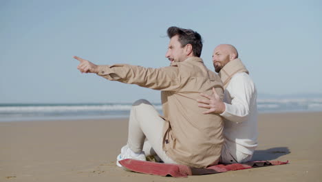 happy gay man sitting on the sandy beach with his partner and pointing finger into distance