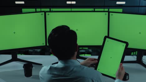 back view of asian man working with tablet and multiple computer monitor with mock up green screen in the office