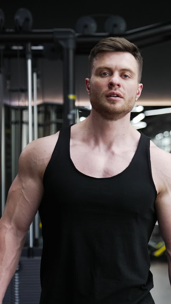 Portrait of a handsome bodybuilder. Young athlete with muscular body walking in modern fitness center on the background of sport simulators. Sportive lifestyle. Vertical video