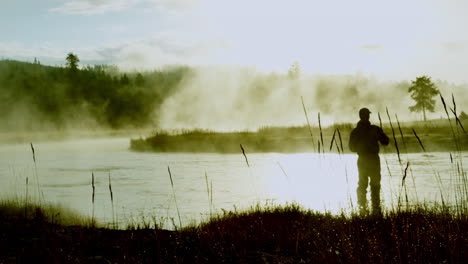 cinematic silhouette shot of a man with fishing rod in his hand at the shore of river illuminated by shiny sunlight and smoke, pov