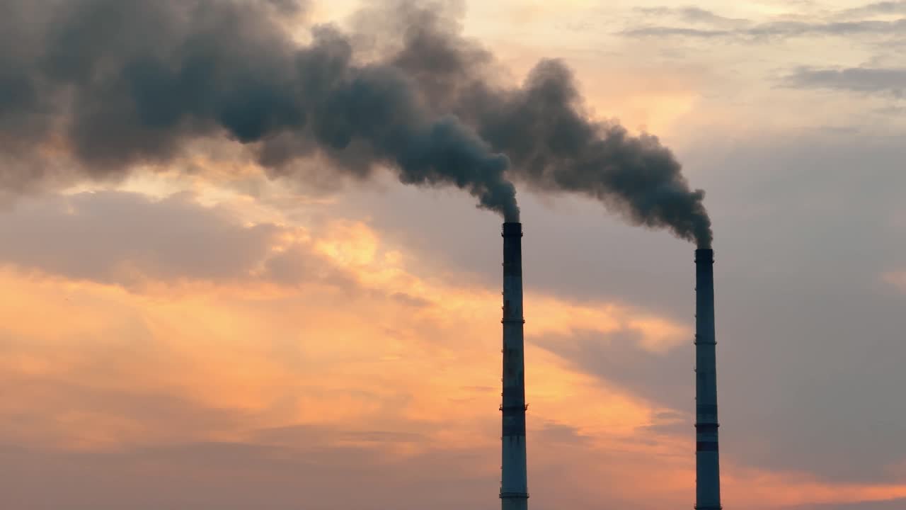 Dei fumaioli emettono fumo nel cielo durante il tramonto in una zona industriale. Due fumaioli rilasciano fumo scuro nel cielo al tramonto in una regione industriale