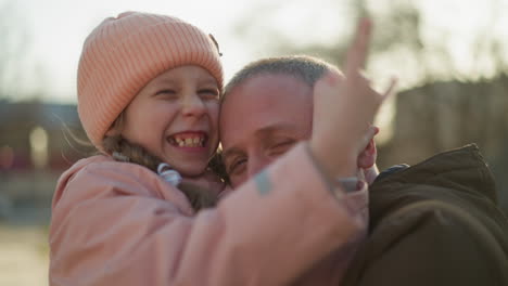 un hombre con una chaqueta marrón lleva a una niña con una gorra rosada y una chaqueta mientras ella hace una señal de mano lúdica con una mano y coloca la otra en su cara. ambos están sonriendo alegremente.