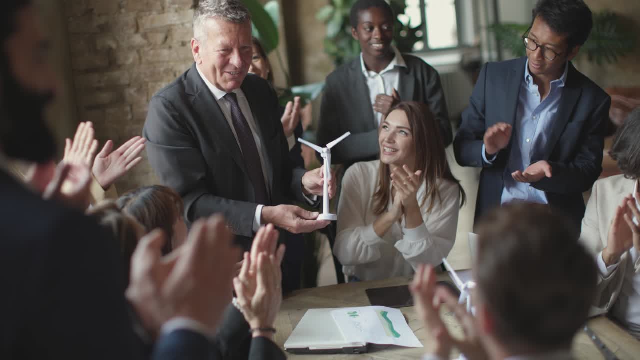 jóvenes compañeros de trabajo aplaudiendo al jefe de negocio con una turbina de molino de viento en la mano - celebración del éxito del proyecto y el trabajo en equipo