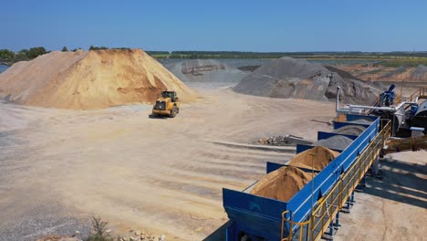 bulldozer transports sand in a bucket. asphalt plant surrounded by piles of crushed stones and gravel. industrial territory of a modern factory outdoors.