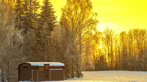 time lapse shot of golden sunset behind snowy fir trees and wooden cabin in foreground