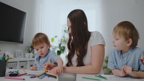 A-young-mother-with-two-children-sitting-at-a-white-table-draws-colored-pencils-on-paper-helping-to-do-homework