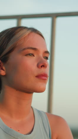 young lady with thoughtful expression as sunlight reflects off her face during dusk, sitting near iron railing, creating moody and reflective atmosphere