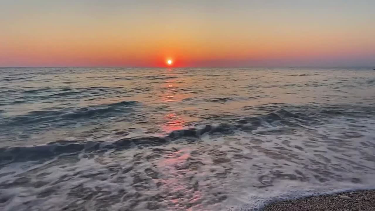 Sunset over calm seaside, waves gently lapping, serene and peaceful coastal view, orange ball of glowing sun lowers to horizon releasing orange yellow glow into sky