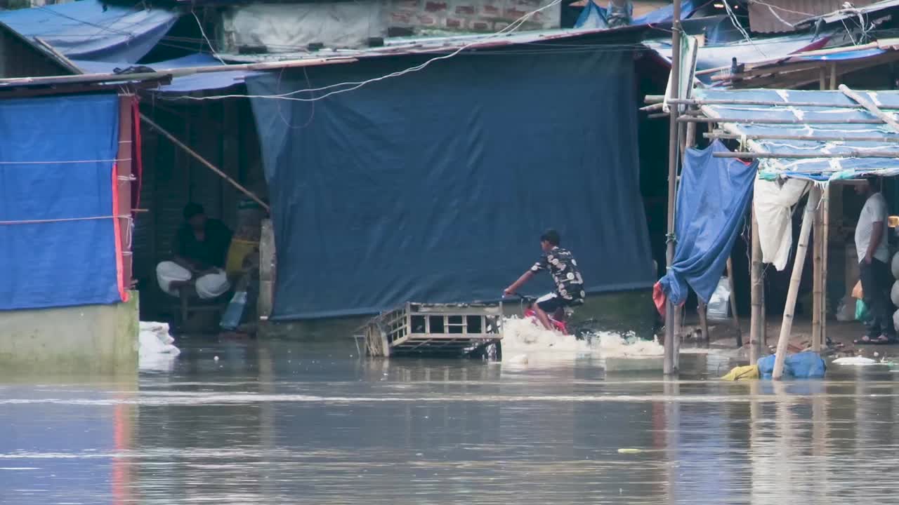 Young Asian boy rides a bicycle in flood water of a slum as muddy water flows in the street