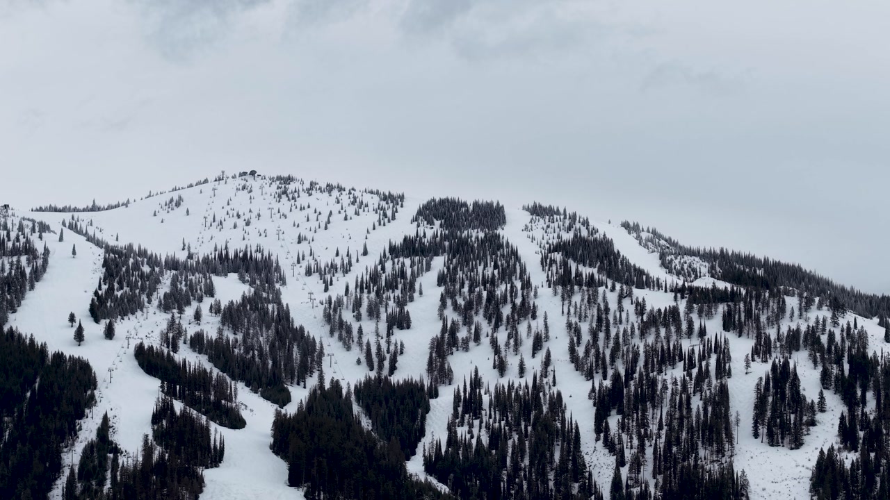 Aerial drone footage capturing the snowy slopes of a mountain ski area near Whitefish, Montana. The camera shows a steep face with groomed runs weaving between evergreen trees under a cloudy winter sk