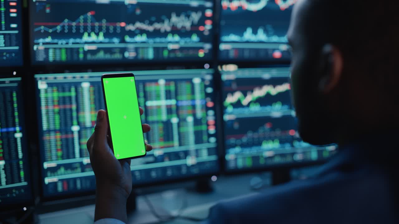 Financial Analyst Using Smartphone with Green Screen Chroma Key Mock Up Template and Working on Multi-Monitor Workstation with Stocks Charts. Businessman Works in Investment Bank in the Evening.