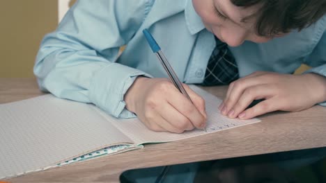 Ext-CU-Tracking-Schoolboy-does-homework-writes-a-pen-in-a-notebook-on-the-table-is-a-tablet