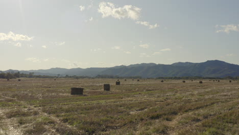 Fly-over-haystacks-in-the-countryside-field-with-mountain-ridge-in-the-background-on-a-bright,-clear,-sunny-day