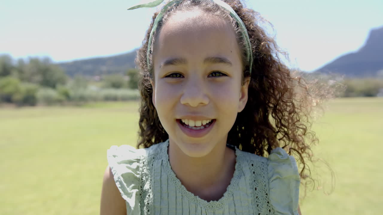 Biracial girl with curly hair smiles outdoors in school, wearing a green dress and headband