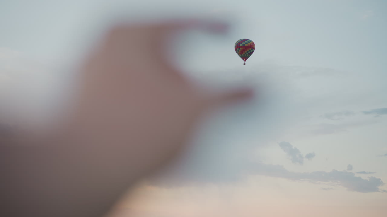 blurred hand waving in foreground framing distant colorful hot air balloon drifting across pastel sky over serene fields with silhouetted trees under soft evening