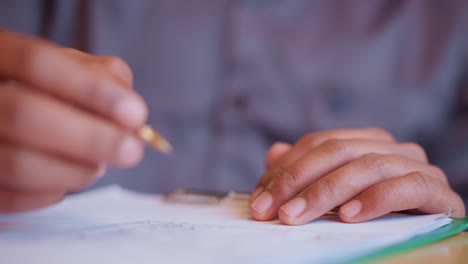 close-up hands holding pen and writing on paper, fingers steady over clipboard, gesture indicates focused activity, likely form filling, studying, or note-taking, shirt in background adds casual setting feel