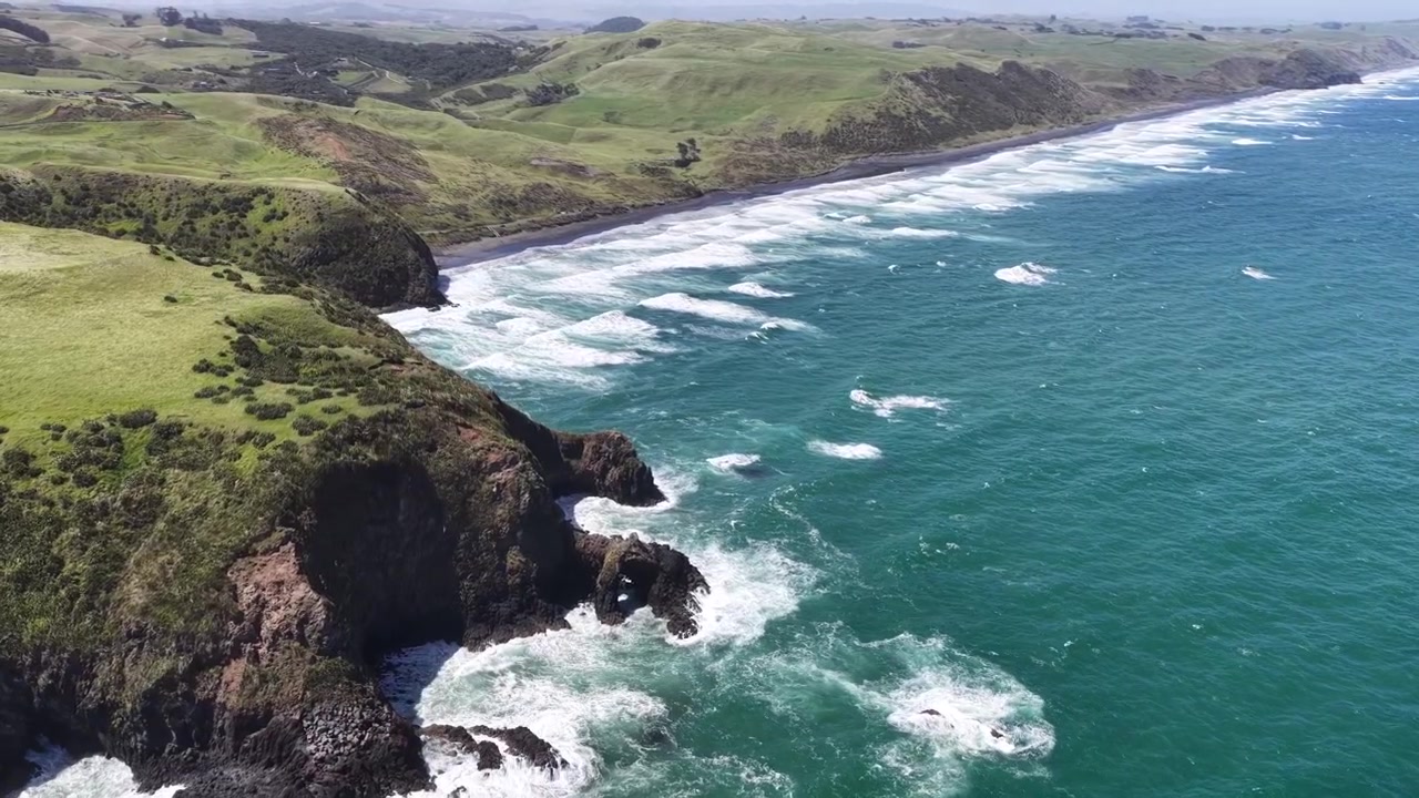 Ripresa ravvicinata con drone su scogliere costiere verdi e onde turchesi che incontrano una spiaggia di sabbia nera, Isola del Nord della Nuova Zelanda, vista paesaggistica mozzafiato.