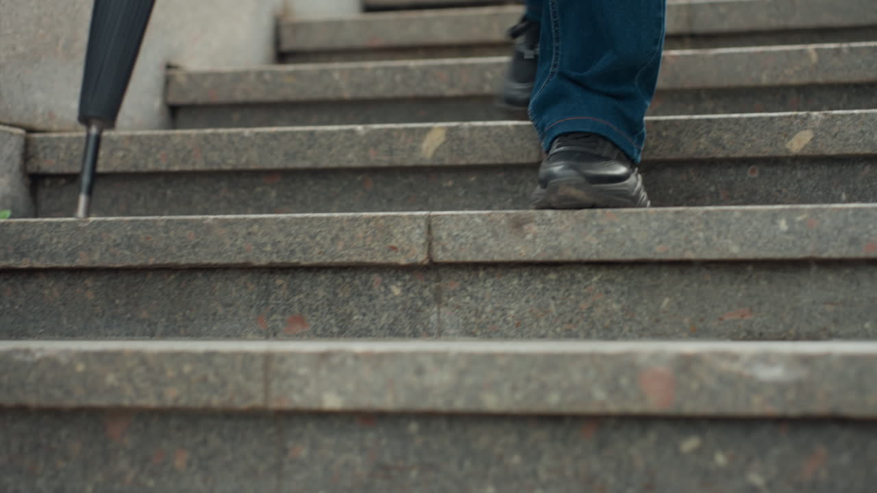 Uomo che scende gradini di pietra con un ombrello in mano, pendolare urbano in blue jeans e scarpe nere che scende con cautela scale di granito bagnate, tenendo la punta dell'ombrello per trovare l'equilibrio, primi piani con scatti dal basso