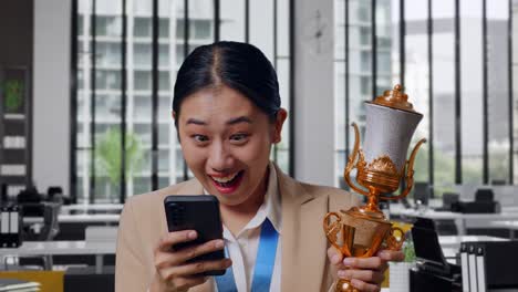 Close Up Of Asian Business Woman In A Suit With A Gold Medal And Trophy Looking At Smartphone Then Saying Wow In The Office