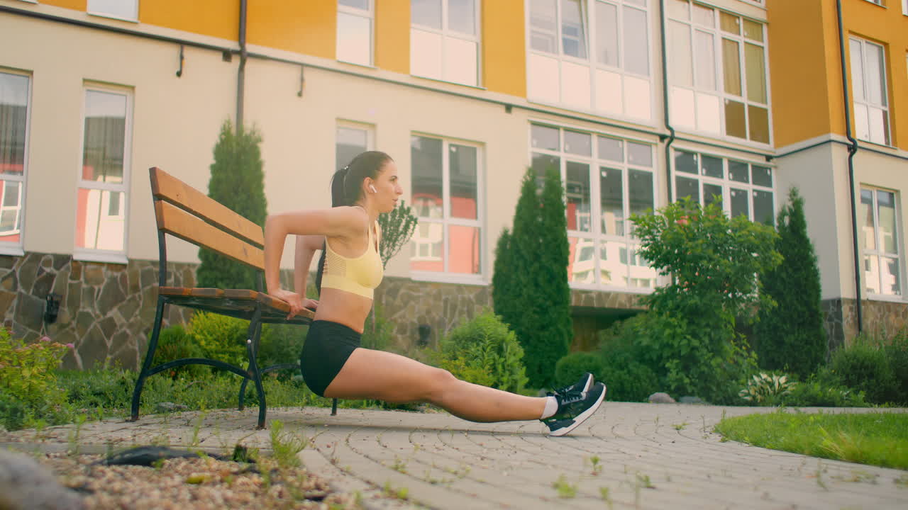 una mujer joven con auriculares realiza flexiones en un banco en un parque de la ciudad en cámara lenta. entrenando a una mujer joven en un banco con el telón de fondo de la ciudad y las casas