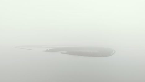 Aproximación-Aérea-Lenta-De-Una-Isla-De-Forma-Ovalada-Cubierta-De-Bancos-Misteriosos-Y-Neblinosos-En-Un-Clima-Diurno-Totalmente-Nublado-Y-Gris-Monótono