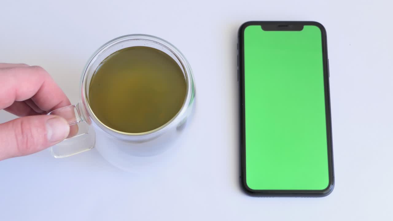 Place for tea drinking with smartphone on white table with green screen. Green tea in a glass cup. Close-up of mobile phone on white table with chroma key, green screen phone and top view