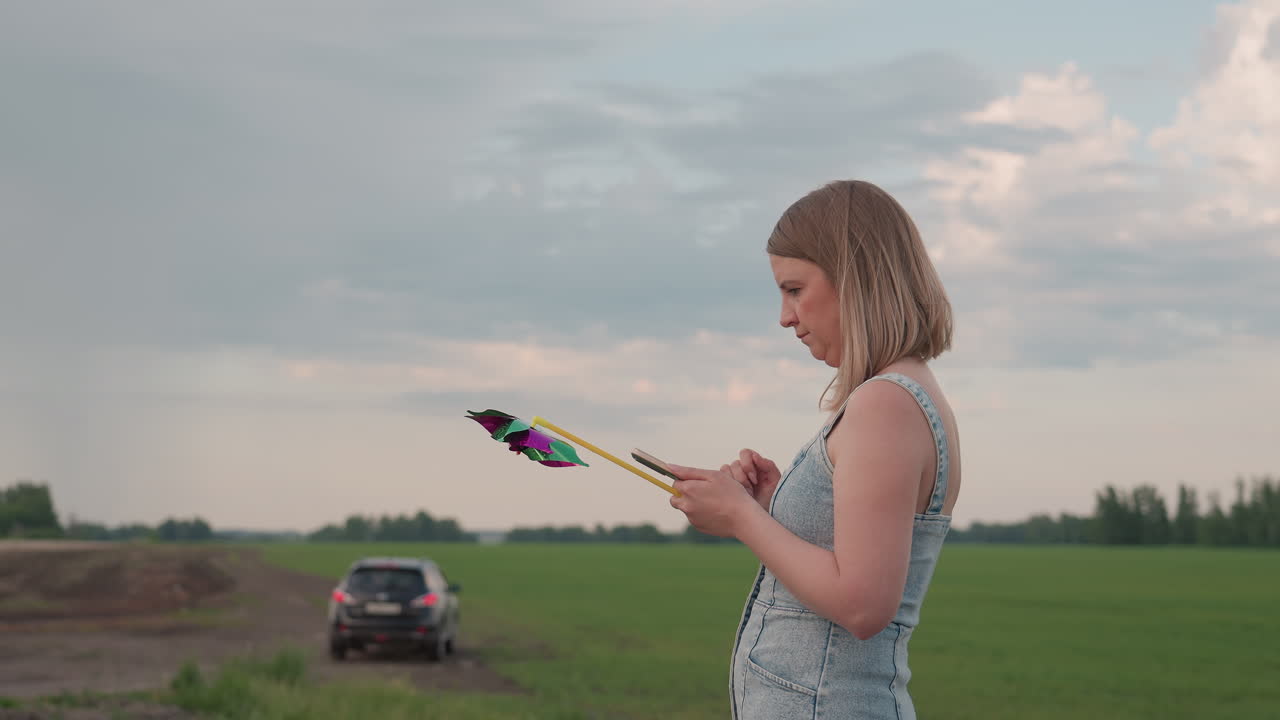 Young woman holding colorful pinwheel glances at smartphone while standing on quiet country road beside green field and distant parked car under blue sky with fluffy clouds beside peaceful farmland
