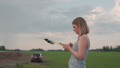 young woman holding colorful pinwheel glances at smartphone while standing on quiet country road beside green field and distant parked car under blue sky with fluffy clouds beside peaceful farmland