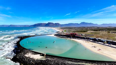 drone view of the shoreline of a cape town beach with a large tidal pool and mountain range in the background on a summers day