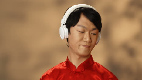 close-up view of cheerful young asian man in red traditional costume wearing headphones and listening to music