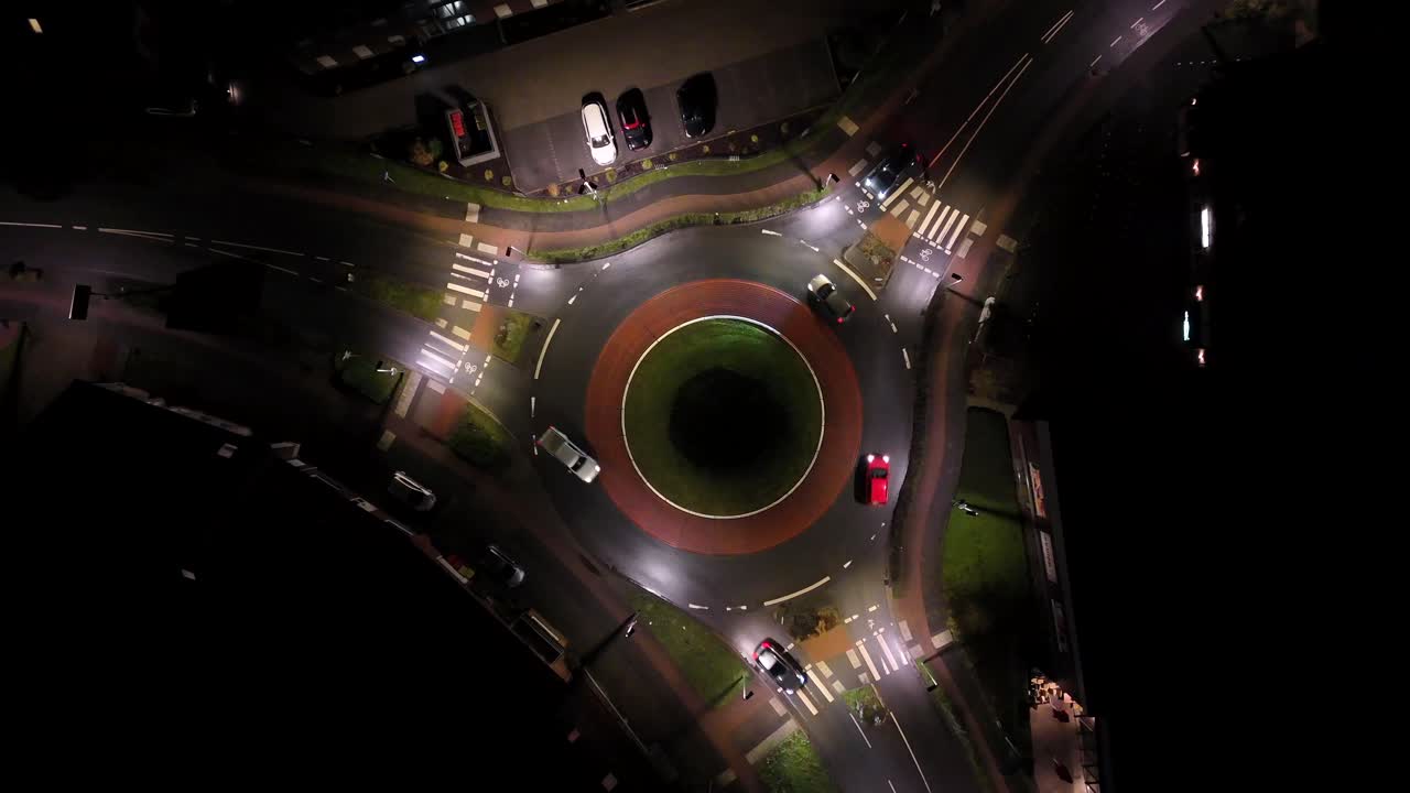 Traffic with cars in roundabout at night. Streetlights illuminating road in american town. Aerial top down landing shot. Walking person on sidewalk.