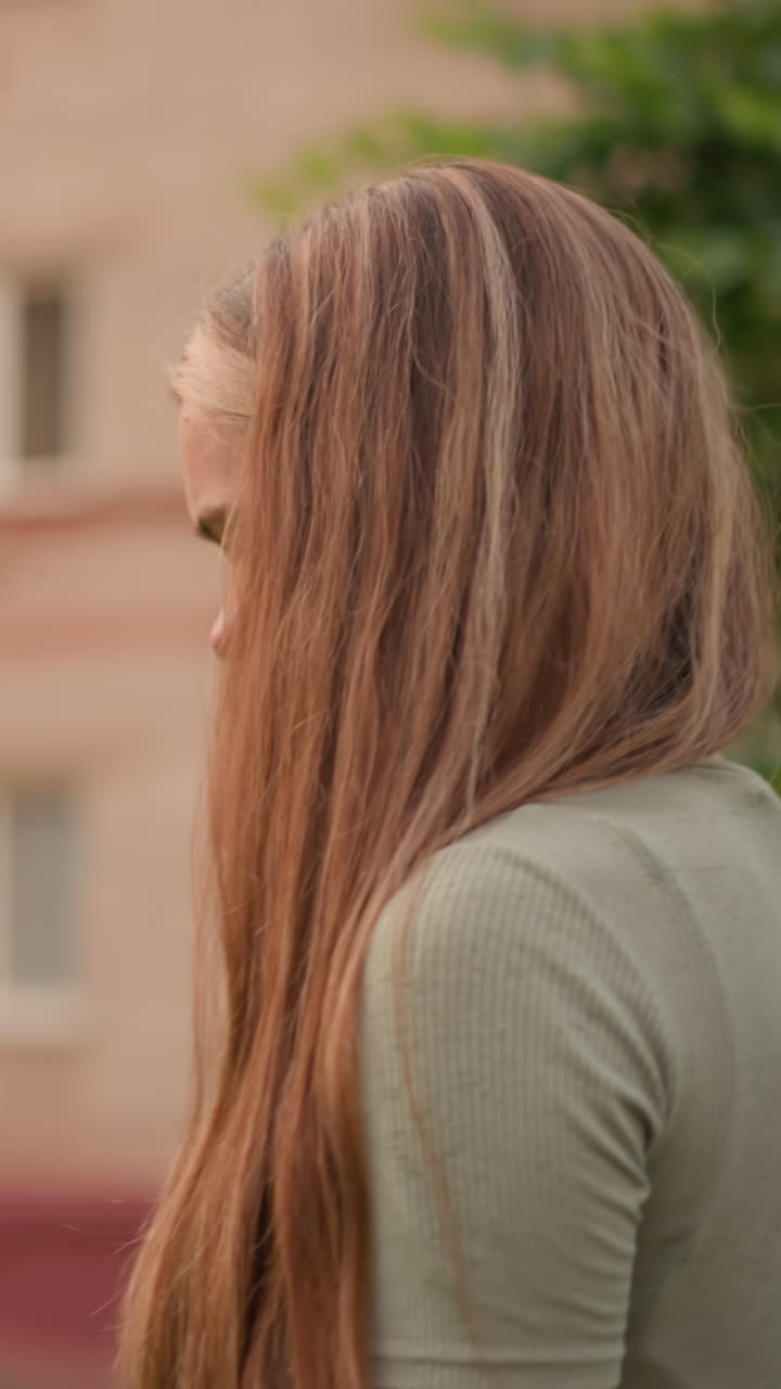 길고 자연스러운 머리카락을 가진 젊은 여성은 건물을 지나가는 동안 사려 깊게 내려다보고, 그녀는 상하고 피곤해 보입니다.