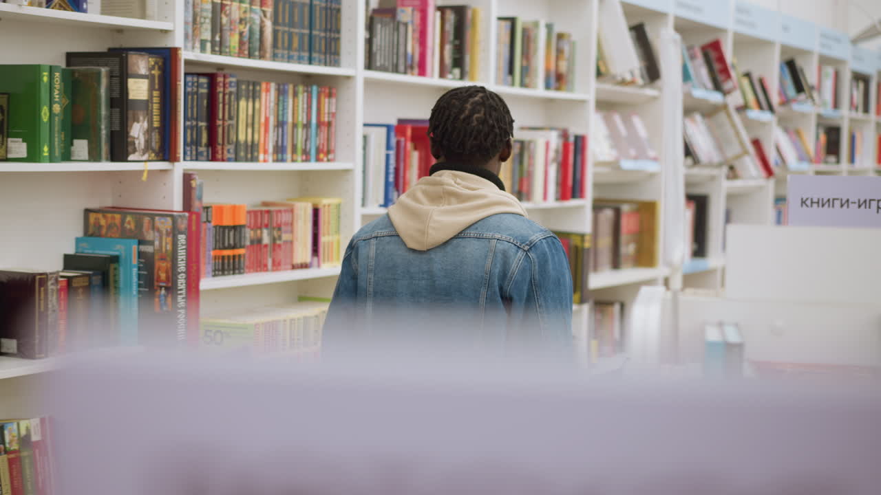 Back view of person in denim jacket with hoodie, standing in front of bookshelves filled with books in library. Focus on individual browsing in a modern, organized bookstore environment