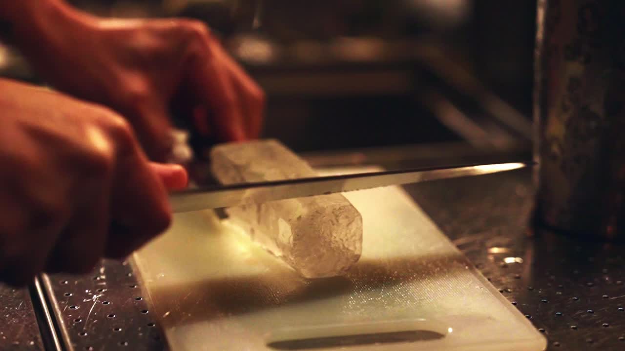 A close-up of a skilled bartender's hands using a sharp knife to precision-carve a crystal-clear block of artisanal ice on a cutting board at bar
