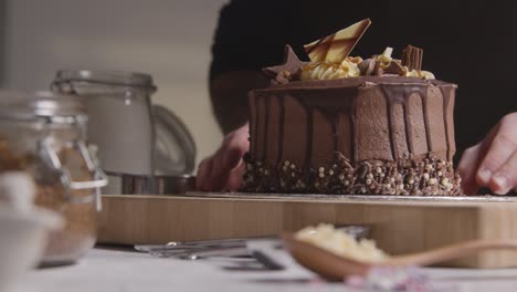 close up of man in kitchen at home putting freshly baked and decorated chocolate celebration cake on work surface