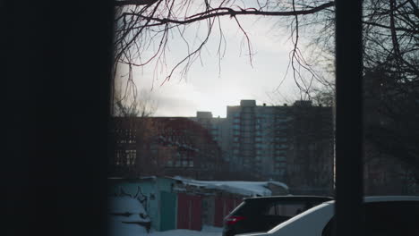 scenic urban view of snow covered area with parked car, dry leafless trees and distant high rise buildings under cold winter sky, soft natural light creating calm peaceful city atmosphere