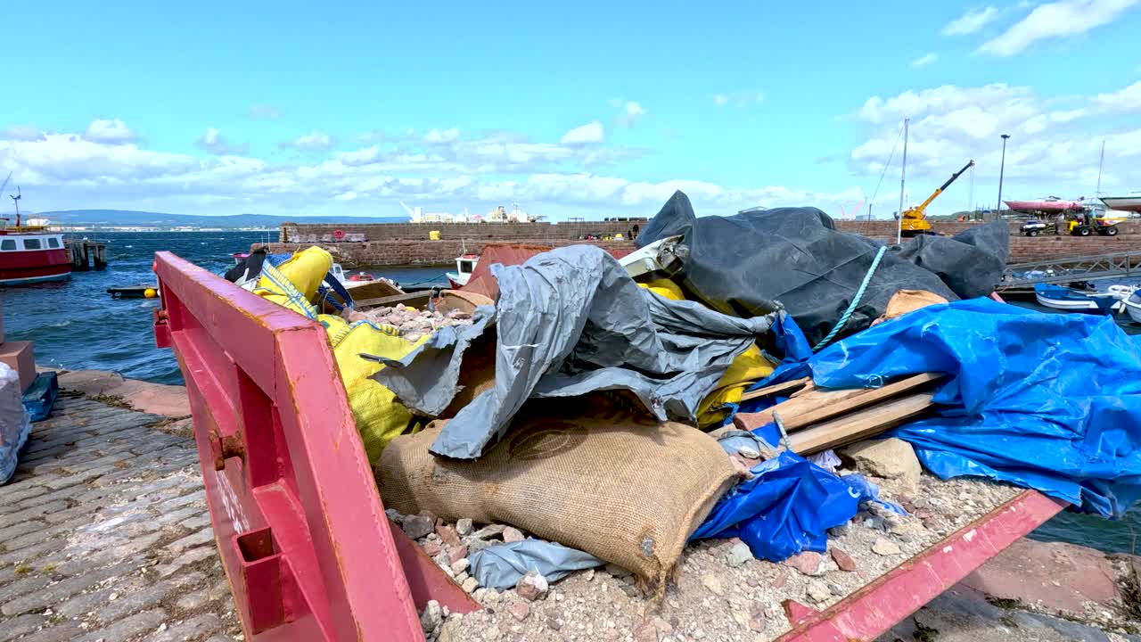 A stationary camera captures a skip filled with plastic, cloth sacks, and debris on a stone pier by the sea under bright daylight