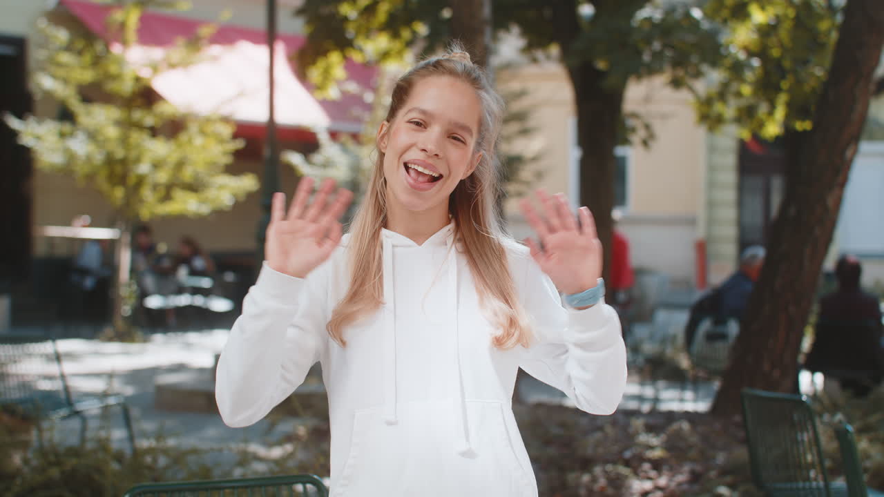 즐거운 젊은 여성 관광객이 카메라를 향해 미소 짓고 손을 흔들며 도시 거리에 서서 안하세요