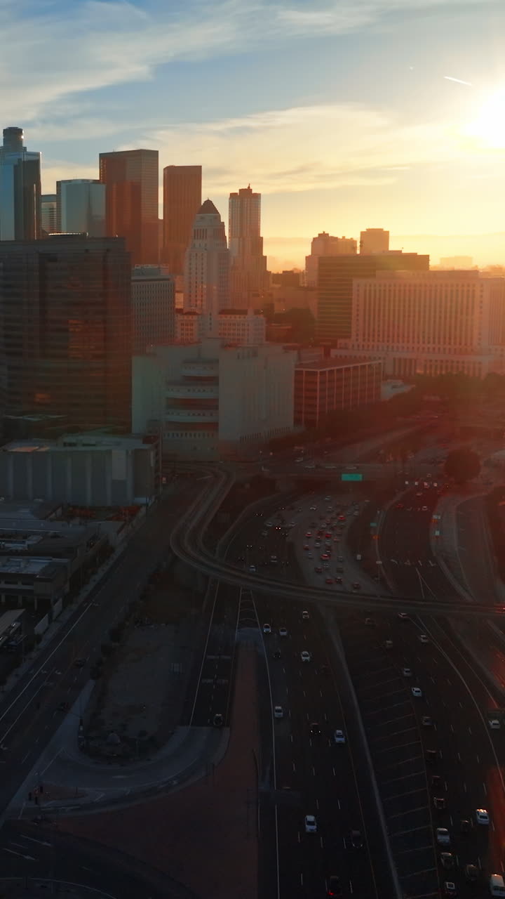 Multiple cars move by the roads and stand on the parking lots. Beautiful downtown skyscrapers of Los Angeles in the rays of setting sun. Vertical video