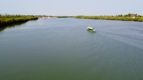 Aerial-drone-shot-of-a-river-in-Gambia-while-a-green-sailing-boat-swimming-forward-with-a-view-of-landscape-in-Kartong-on-sunny-summer-day