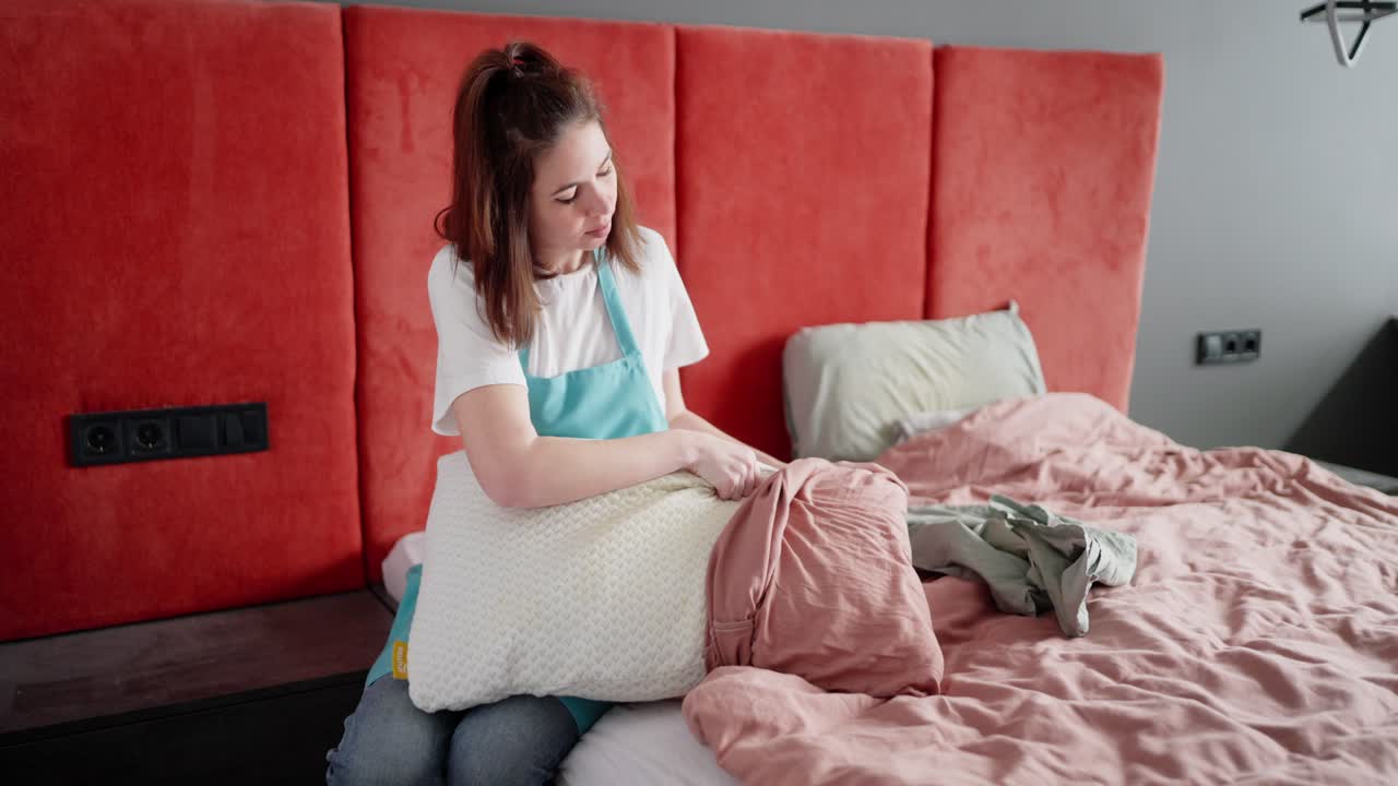 una confiada dama de limpieza morena en una camiseta blanca y un delantal azul mete una almohada en una funda de almohada gris mientras limpia el dormitorio en un apartamento moderno