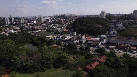 Drone-moving-backwards,-in-the-background-the-urban-landscape-of-São-Paulo-city-and-in-the-first-plan-a-green-area-with-some-trees-and-a-lake