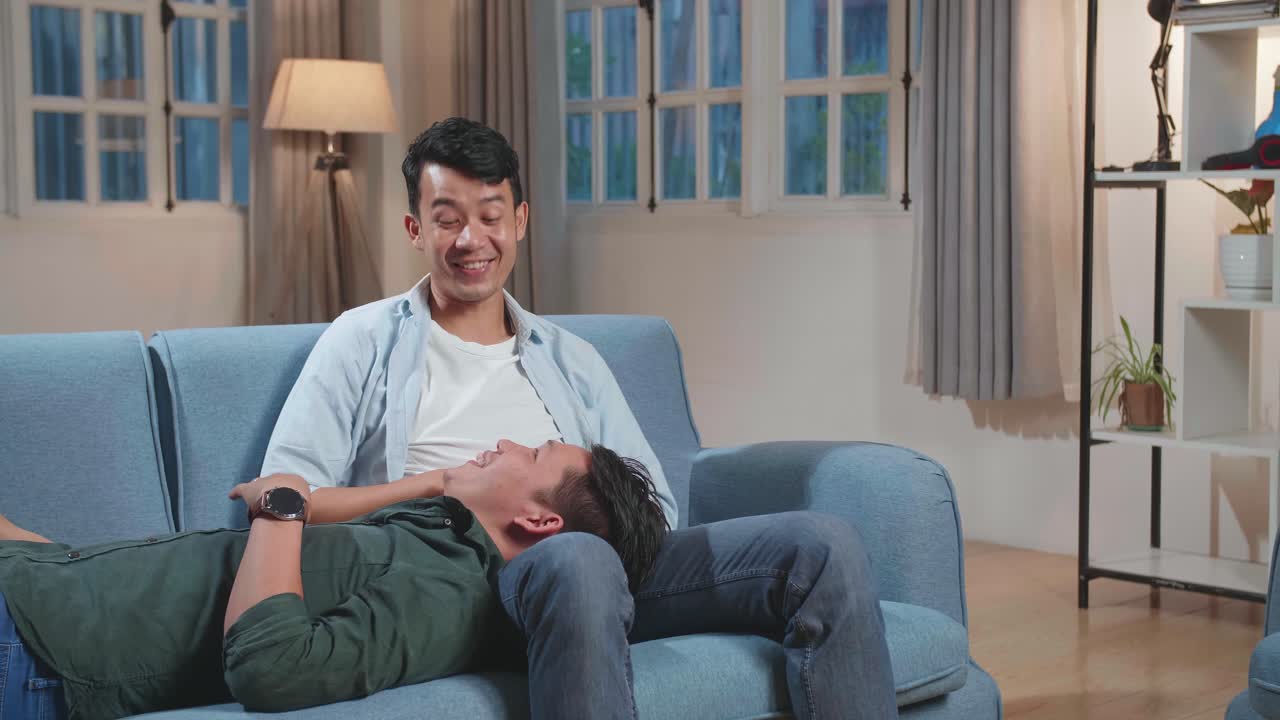Young Asian Gay Couple Spend Time At Home. Two Happy Man In Love Lie Down On Sofa And Cuddle In Casual Clothes. Cute Lgbt Relationship Content