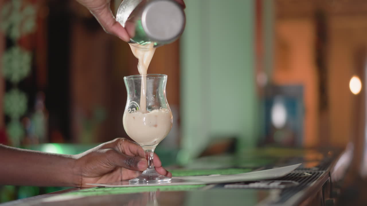 Bartender pouring creamy drink from shaker into glass on bar counter, focused on hands, with blurred background of bar and bar tools