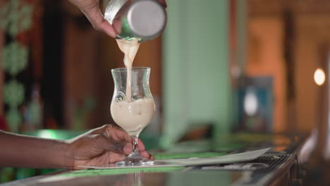 bartender pouring creamy drink from shaker into glass on bar counter, focused on hands, with blurred background of bar and bar tools