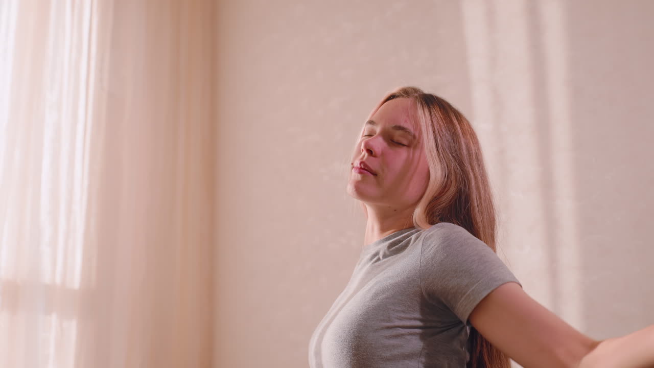 Side view of young girl gently arching back with hands on waist, stretching muscles in morning light while seated on couch, long hair falling behind, sunlight casting soft glow on wall