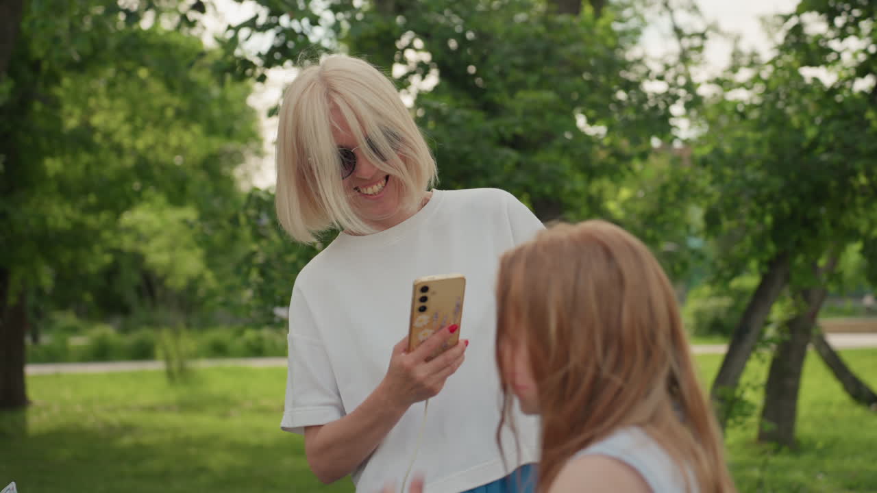 excited mom in casual summer outfit taking pictures of toddler playing near fountain in green park, joyful interaction captured on phone, hair blowing, happy family moment